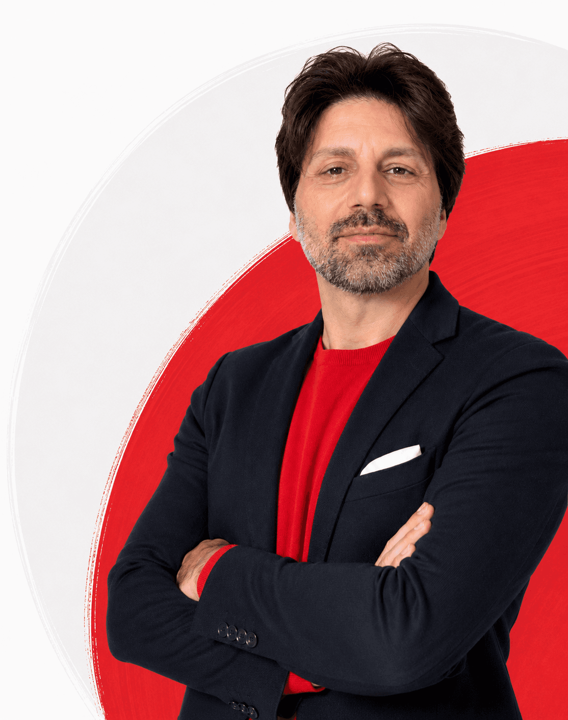 Uomo con barba brizzolata, giacca scura, maglia rossa, braccia incrociate su sfondo bianco e rosso.