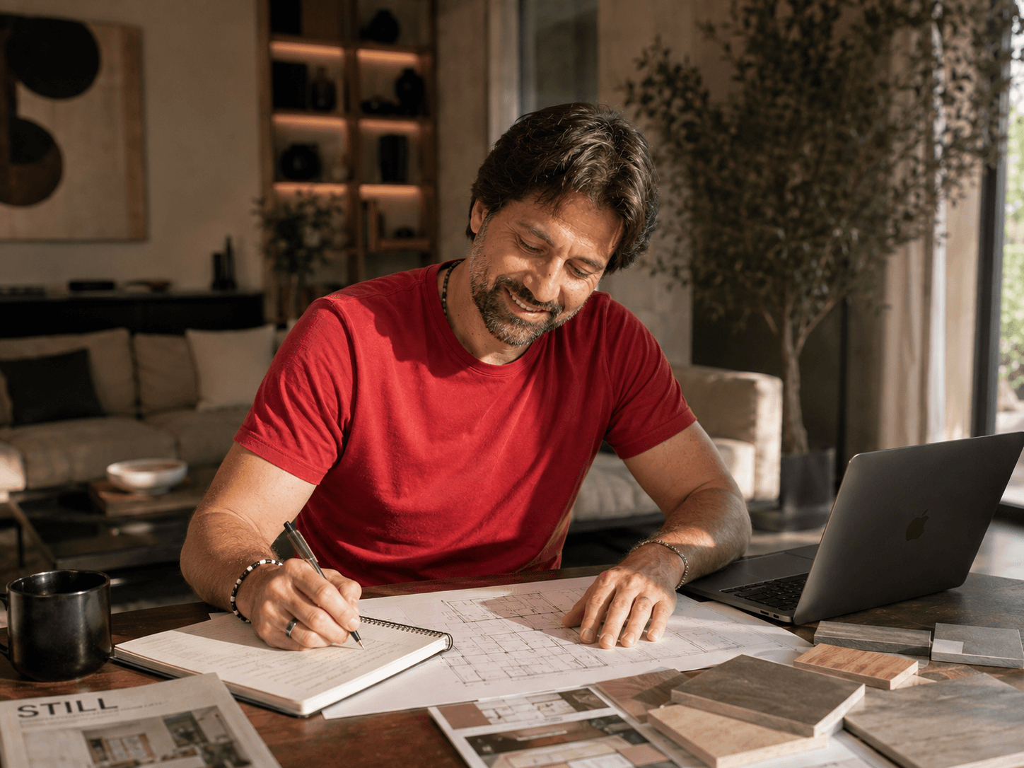 Uomo sorridente in maglietta rossa lavora a una scrivania con planimetrie, taccuino e laptop.