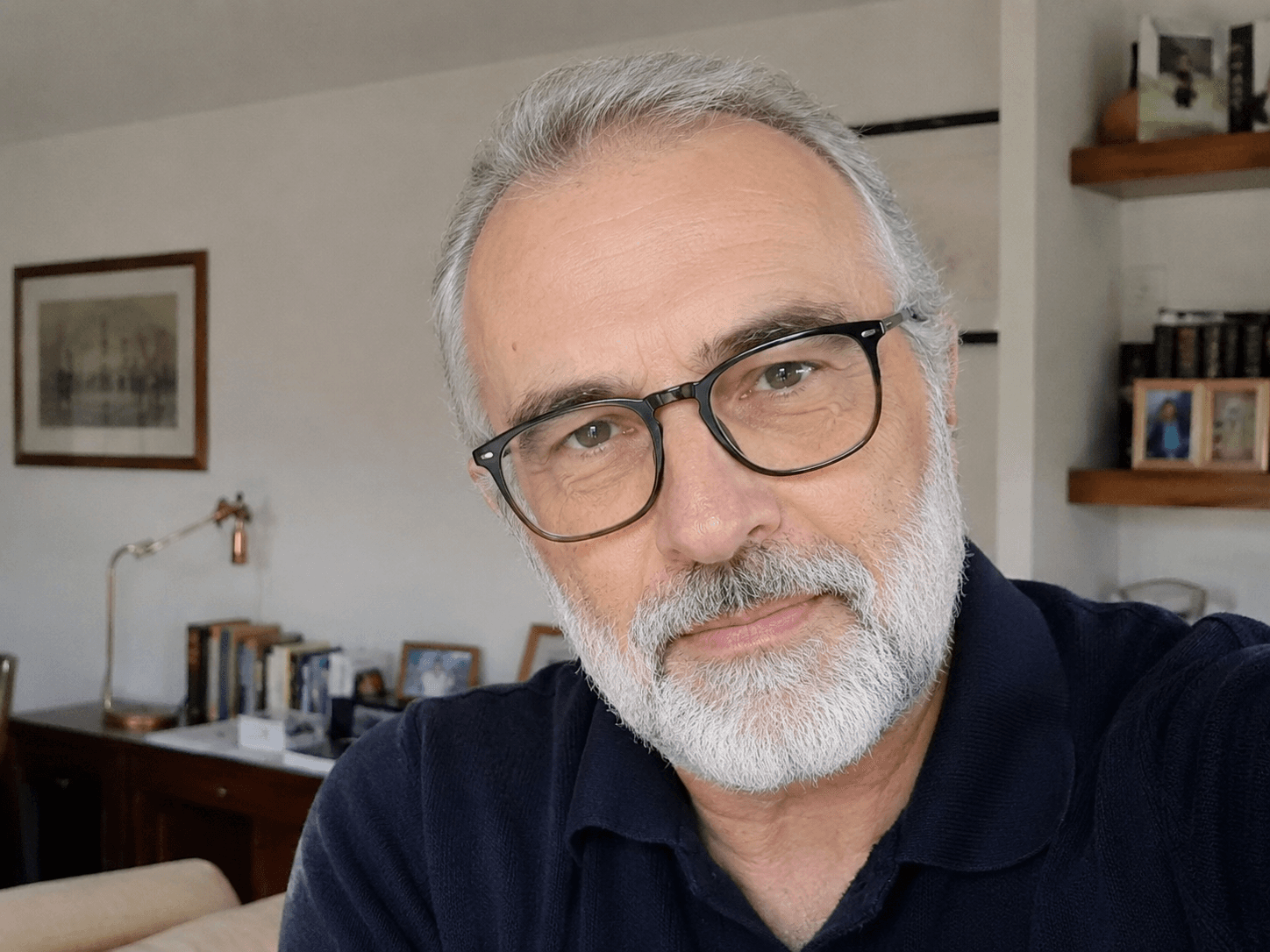 Uomo con barba grigia e occhiali neri indossa una polo blu scuro in un ufficio.