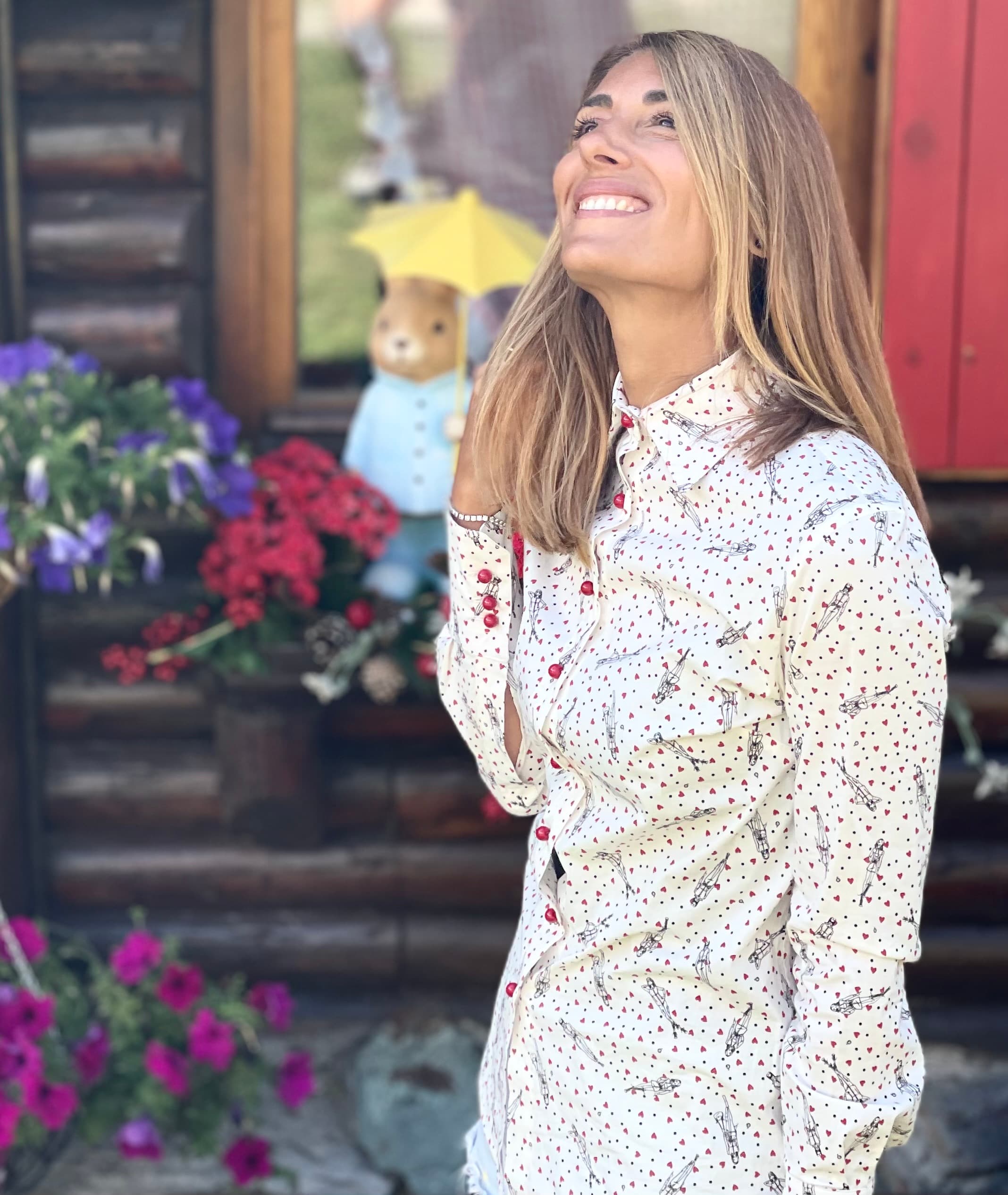Donna sorridente con camicia a fantasia guarda in alto davanti a una baita fiorita.